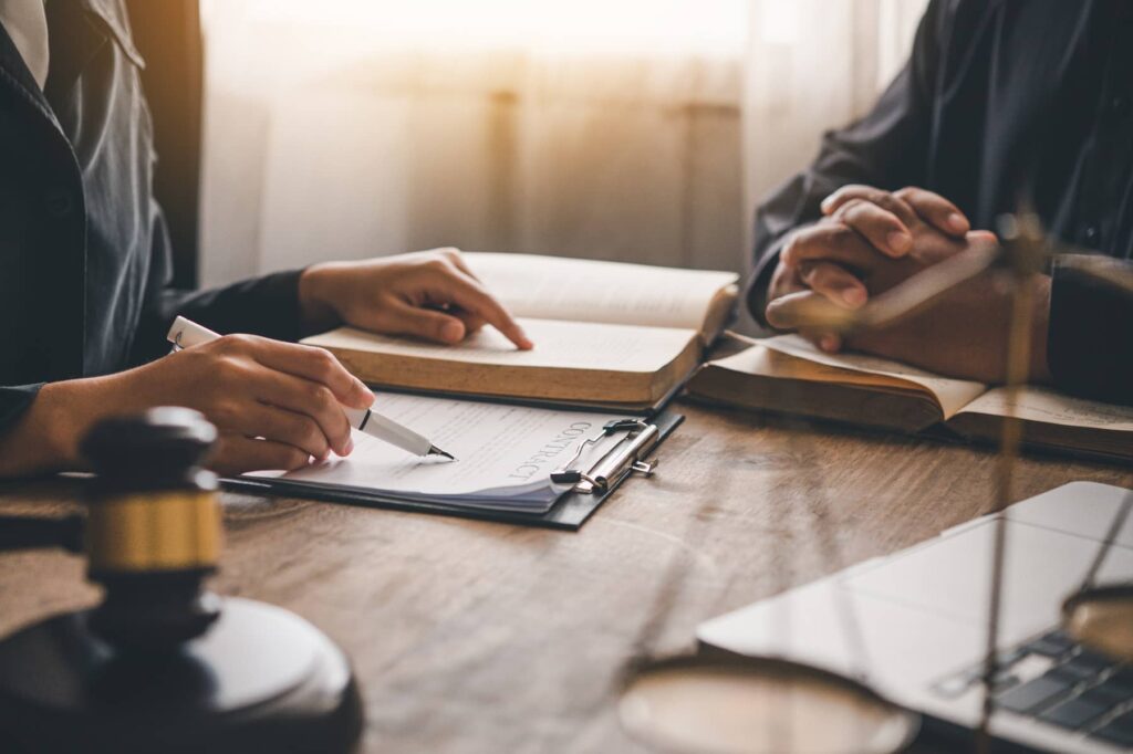 Two people sit at a desk with legal documents, books, a gavel, and a laptop; one person is writing on a contract with a pen.